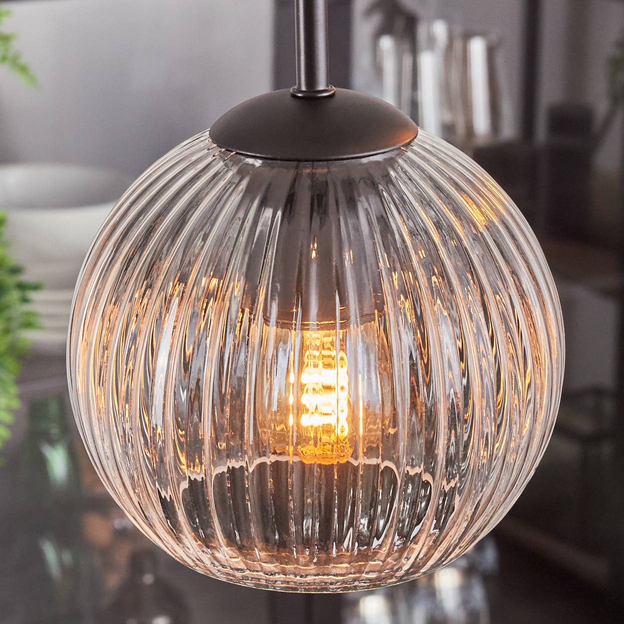 Eine klare, gerippte Glas-Pendelleuchte mit warmer Glühbirne. Dunkle Metallbeschläge. Verschwommener Hintergrund zeigt eine Küchenumgebung.