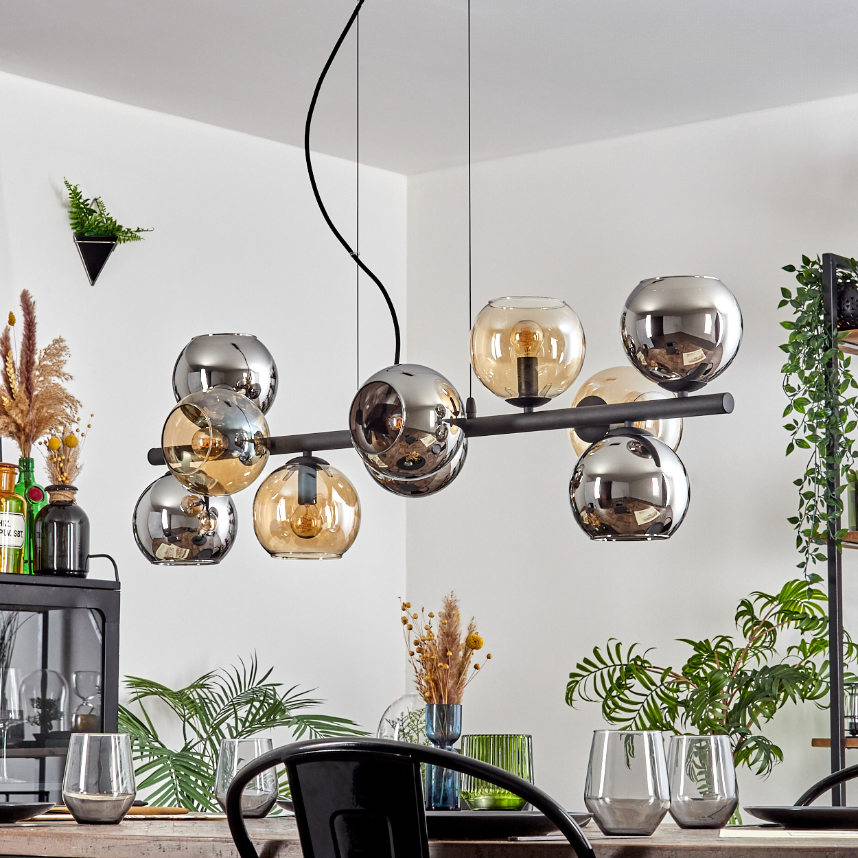 Ein modernes Esszimmer mit einem schwarzen Kronleuchter mit Glaskugeln, Tisch, Stühlen, Pflanzen und Glaswaren. Die Leuchten haben silberne und bernsteinfarbene Kugeln.