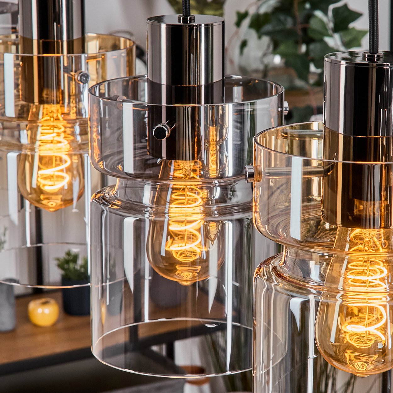 Nahaufnahme von drei modernen Pendelleuchten. Jede hat einen Glasschirm und eine warme, leuchtende Glühbirne. Glänzende Metallakzente reflektieren Licht.