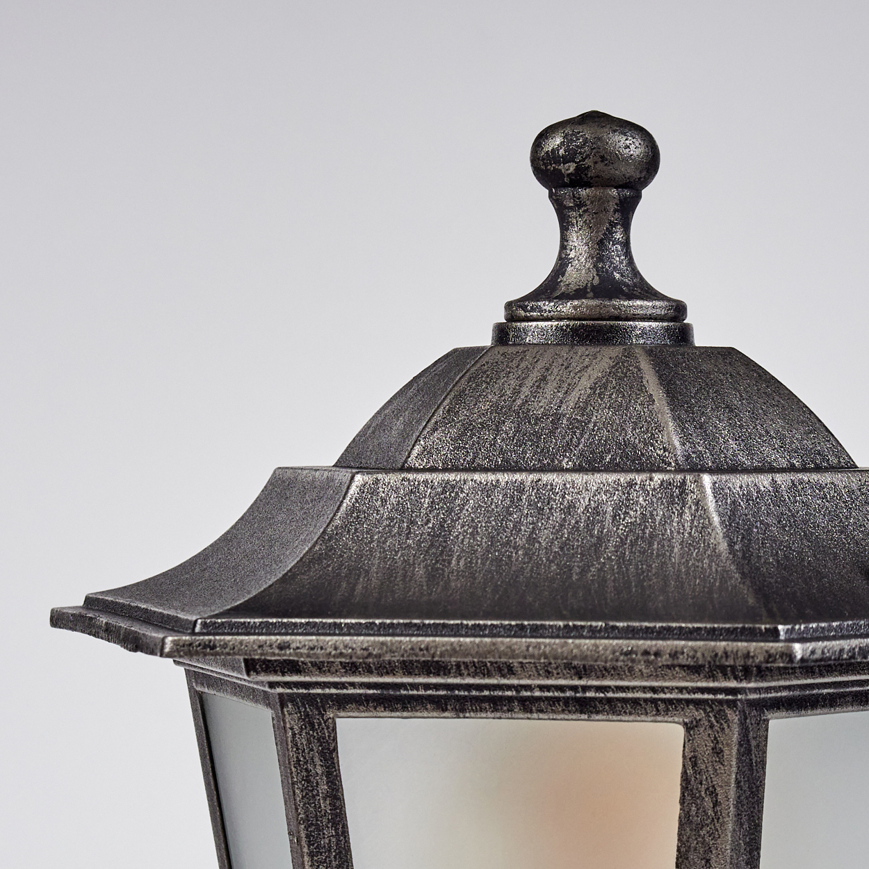 Nahaufnahme einer dekorativen Außenlampe. Sie hat ein metallisches Finish mit einer Kappe und einem geometrischen Design. Der Hintergrund ist weiß.