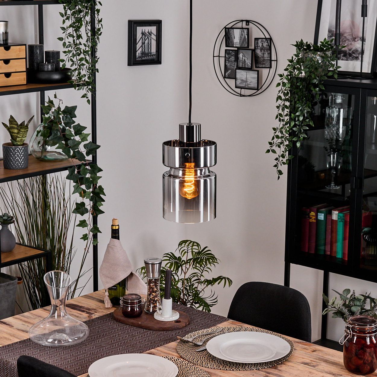 Ein Esstisch mit Tellern, Besteck und einer Hängelampe. Regale mit Pflanzen und Dekor sind im Hintergrund. Ein Glas mit Essen steht auf dem Tisch.