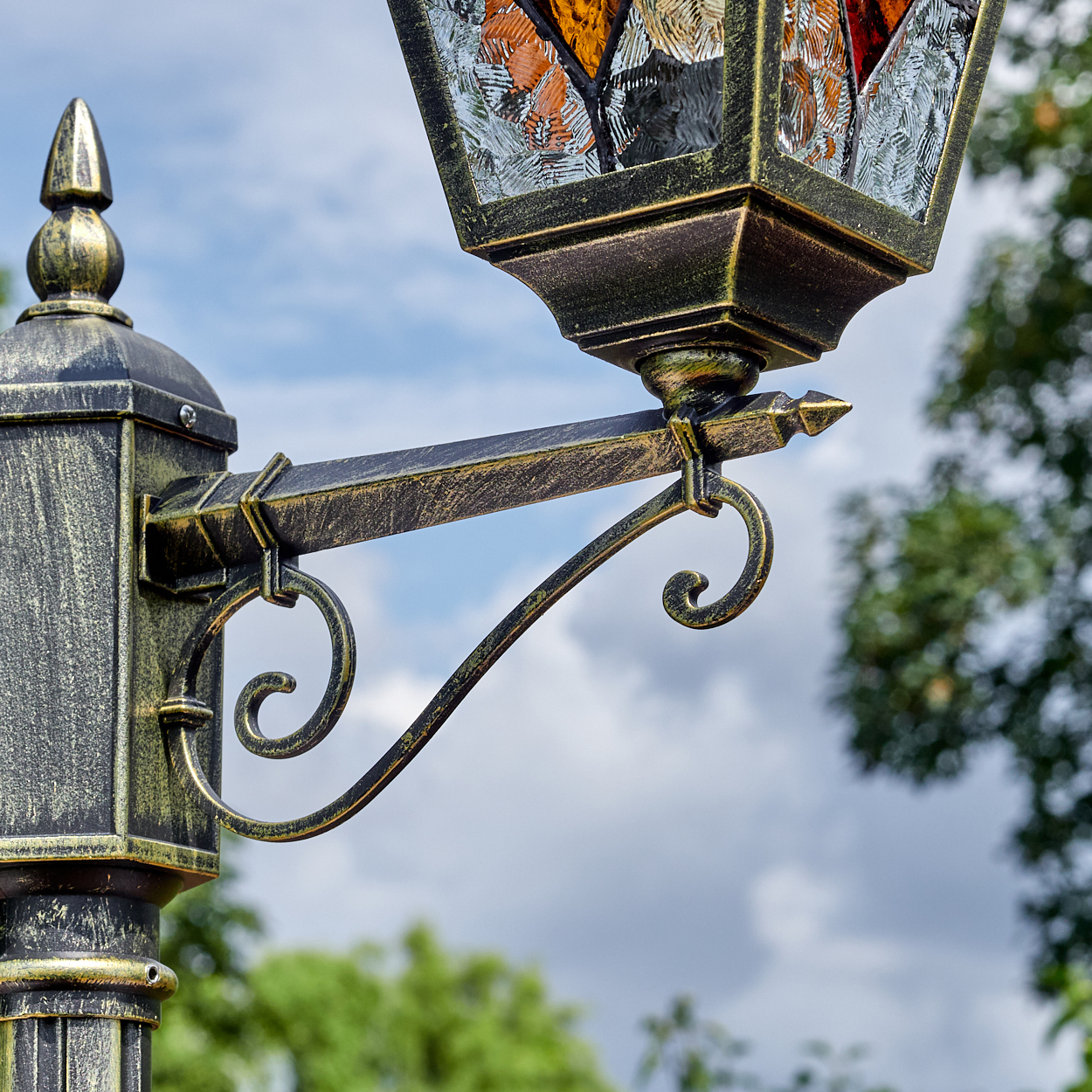 Nahaufnahme einer dekorativen Laterne mit Buntglas. Die Stange ist schwarz und gold mit Himmel und Bäumen im Hintergrund.