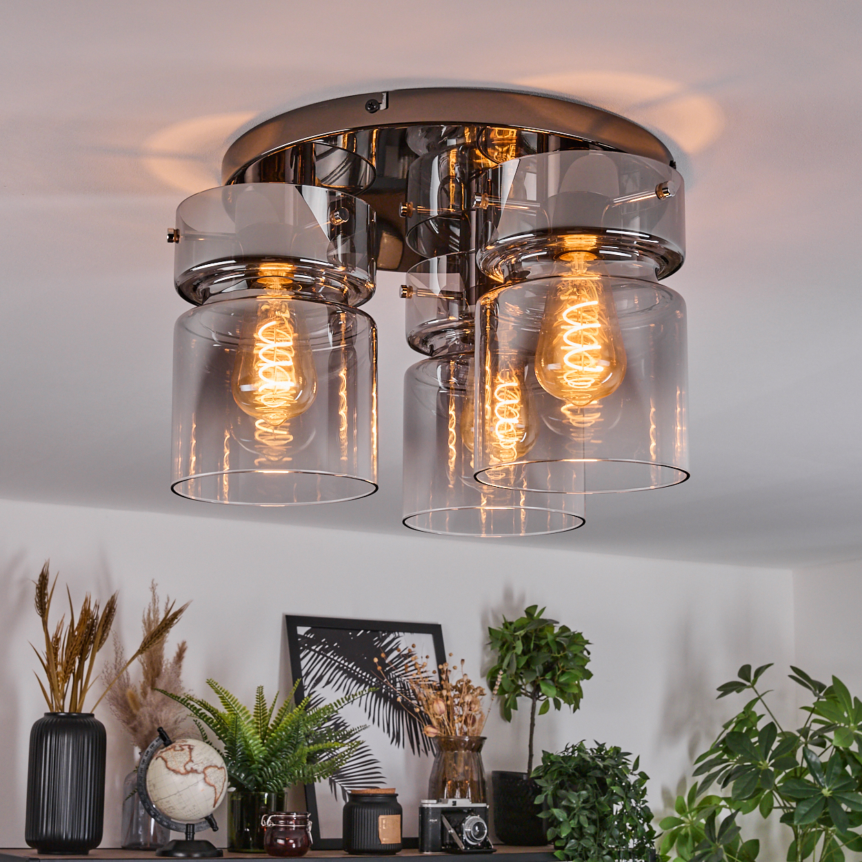 Eine moderne Deckenlampe mit vier Glühbirnen. Darunter ein Regal mit Pflanzen, einem Globus, einem Foto und anderen dekorativen Gegenständen.