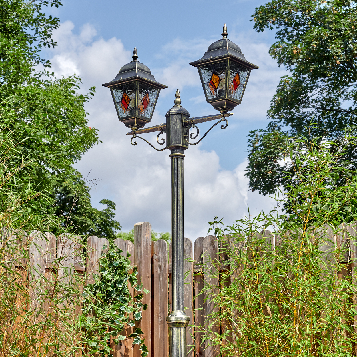 Eine verzierte Laterne mit zwei Lampen. Im Hintergrund ein Holzzaun. Grüne Bäume und Bambus umgeben die Laterne unter blauem Himmel.