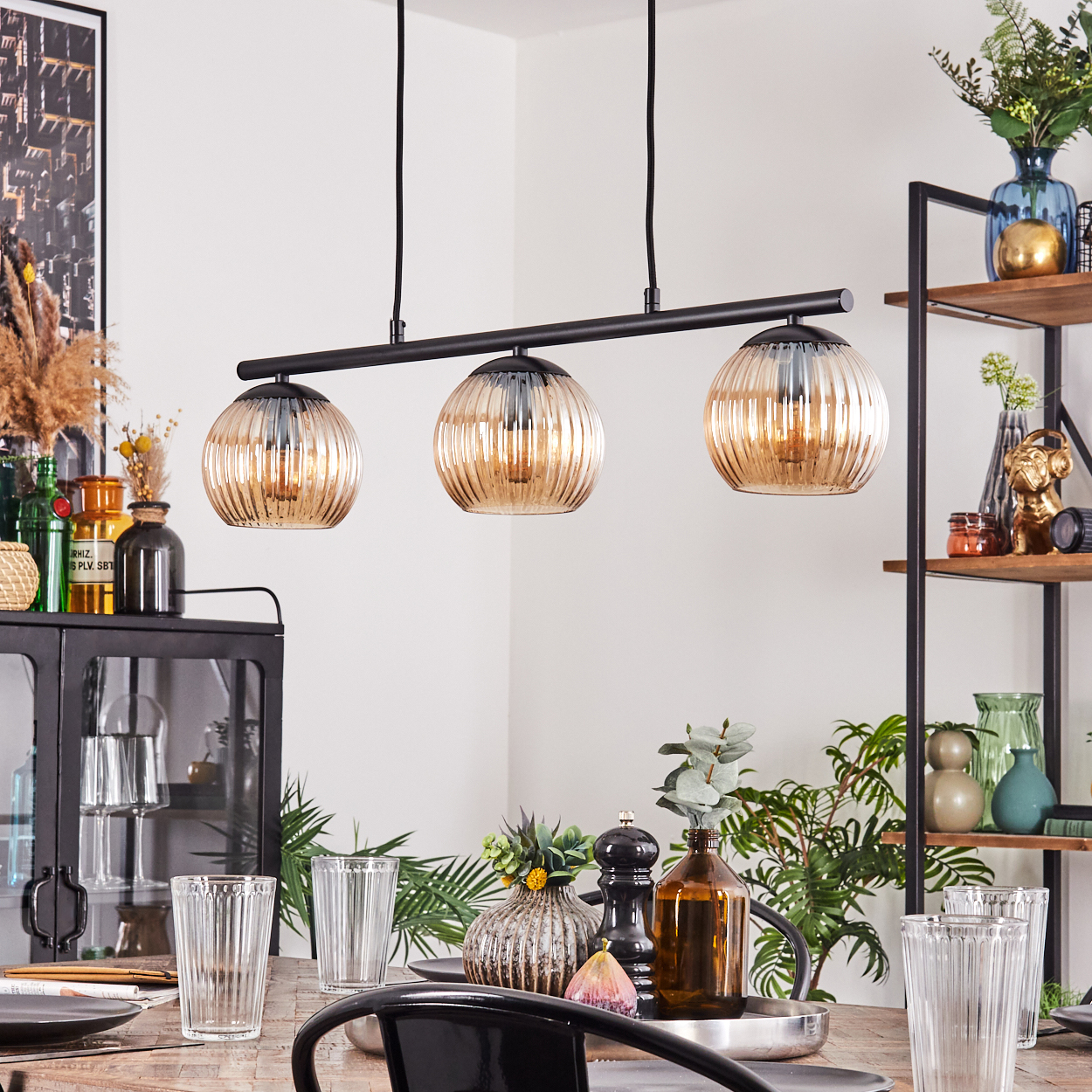 Esszimmer mit gedecktem Tisch, Dreilampen-Pendelleuchte und Dekorationsartikeln auf Regalen.