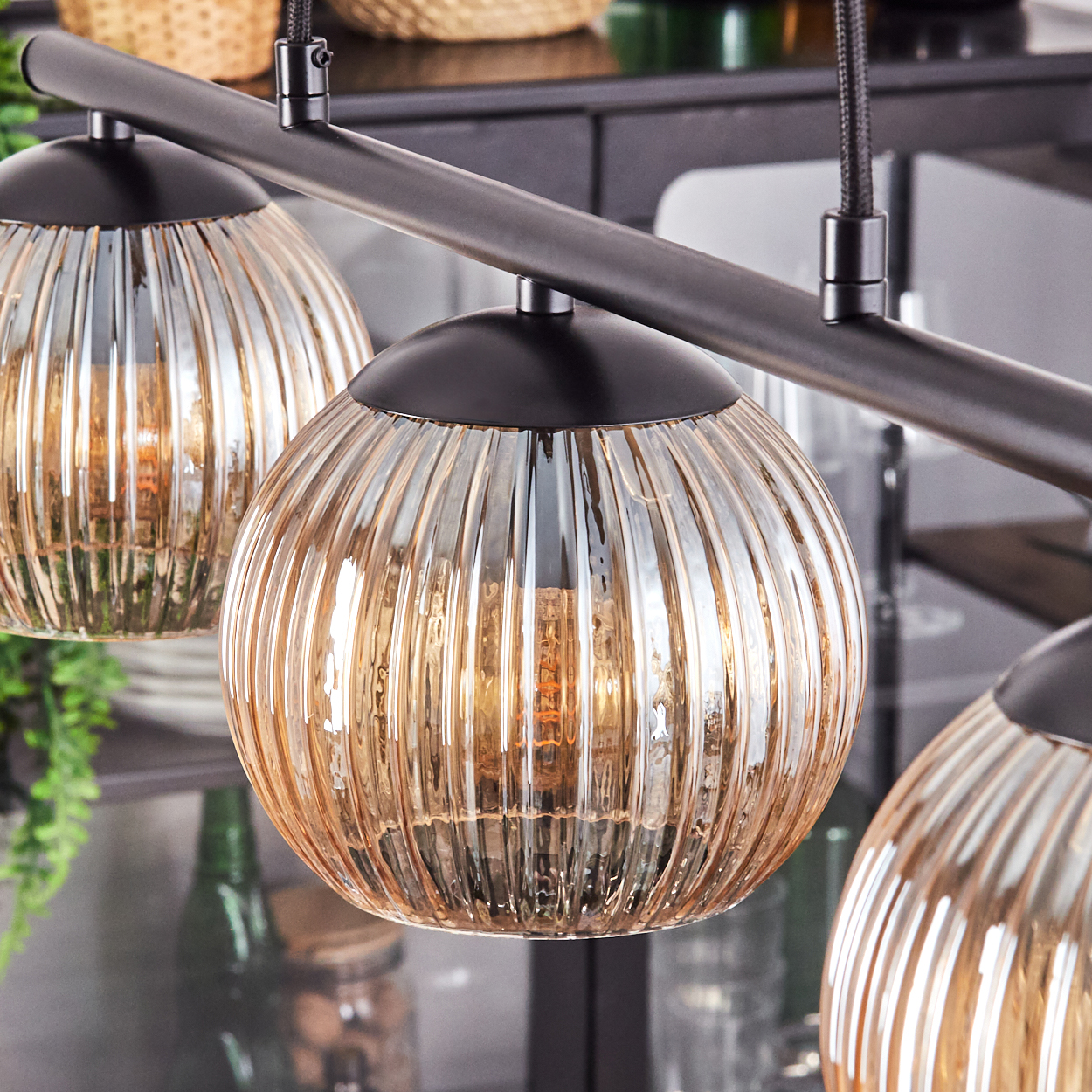 Eine moderne Pendelleuchte mit drei kugelartigen, gerippten Glasschirmen, die an einer dunklen Metallstange hängen. Der Hintergrund ist unscharf.