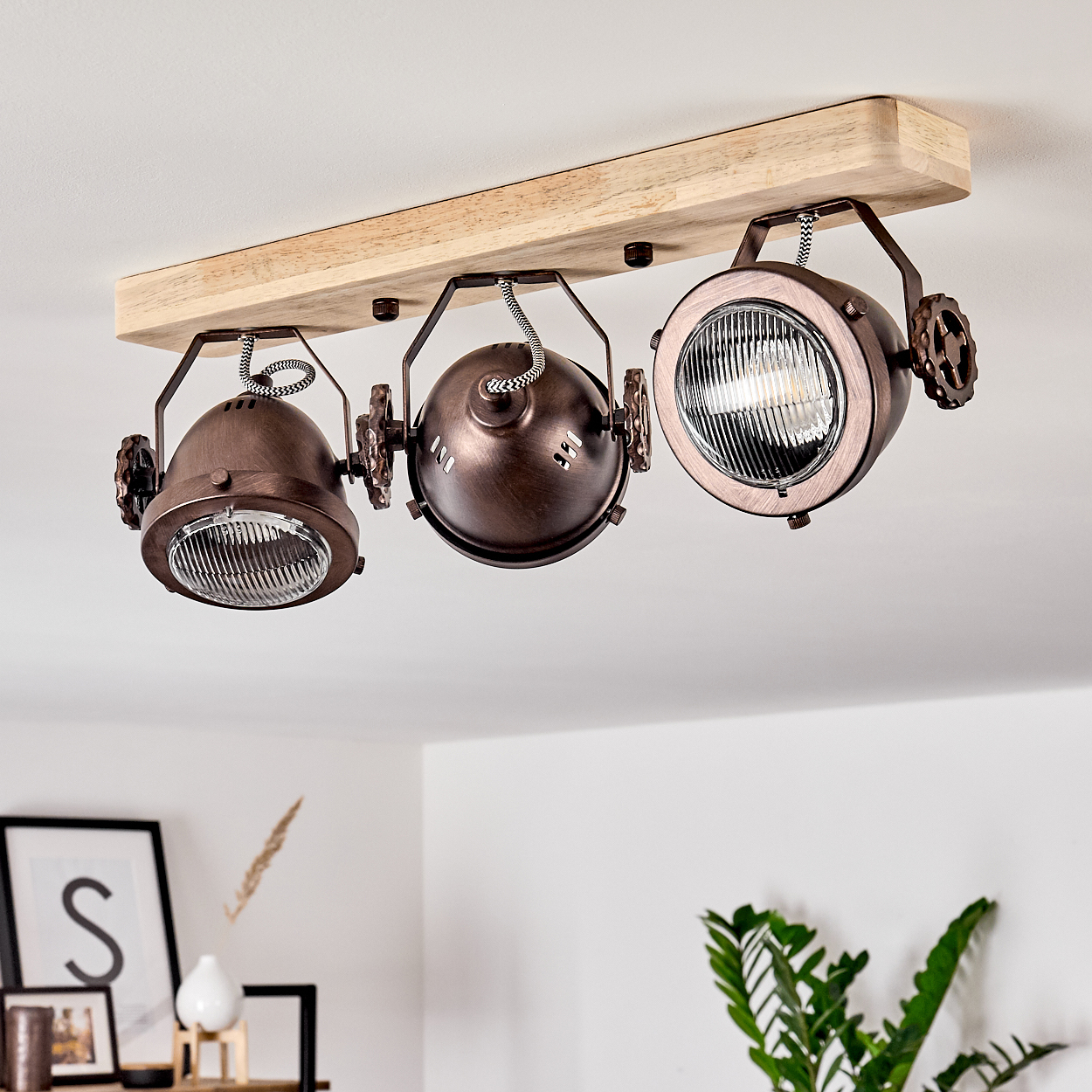 Drei bronzefarbene Strahler an einem Holzbalken. Der Hintergrund ist eine weiße Decke und Wände mit etwas Dekoration.