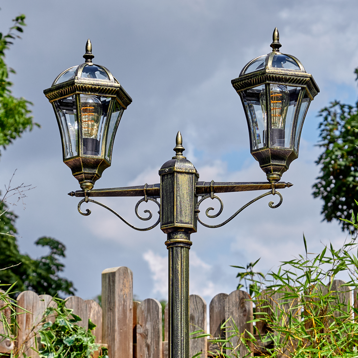 Eine dekorative bronzefarbene Außenlampe mit zwei Lichtern. Sie steht vor einem Holzzaun, mit bewölktem Himmel im Hintergrund.