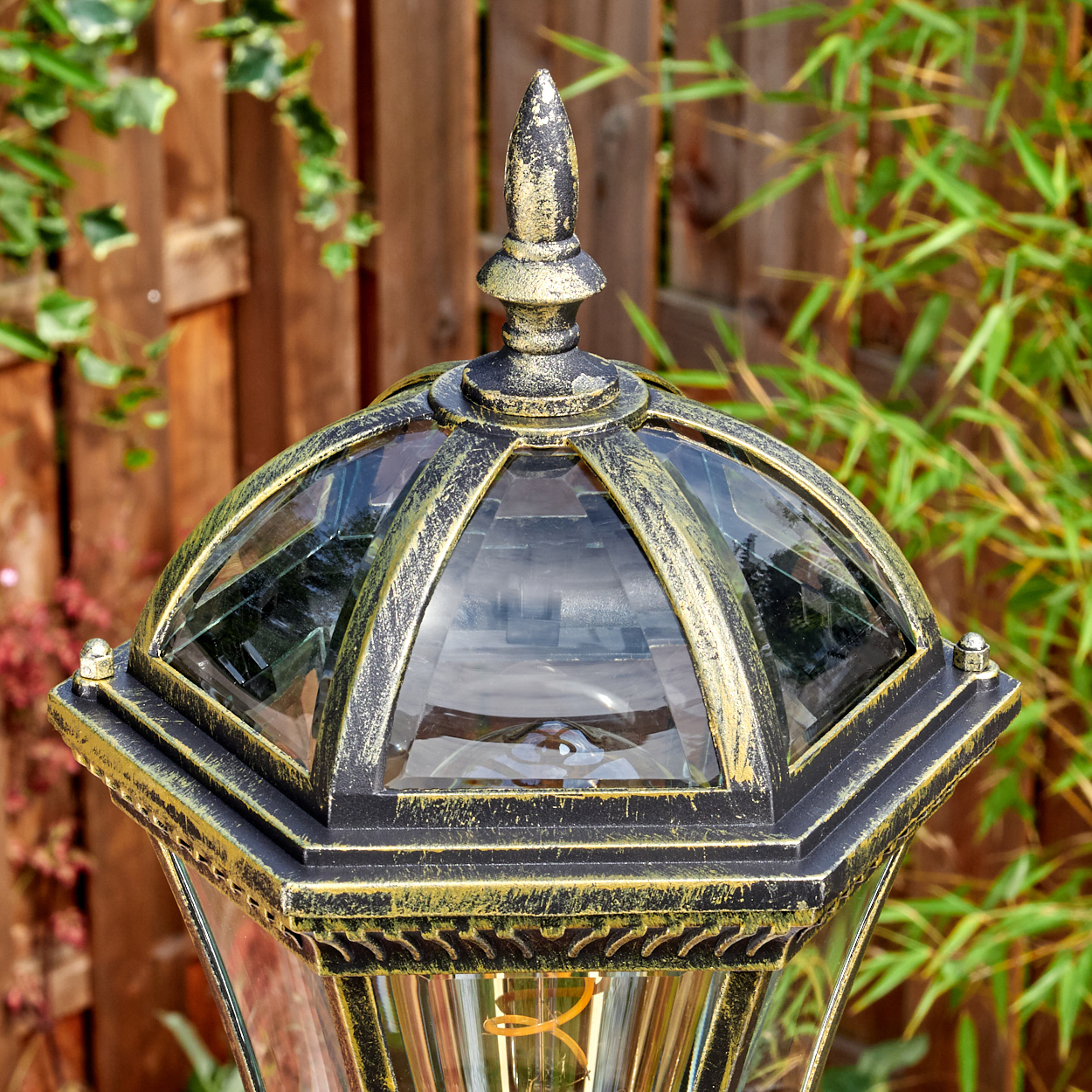 Eine dekorative Außenlampe mit Glaspaneelen und einem gold-schwarzen Rahmen. Sie hat eine spitze Spitze. Ein Holzzaun und grüne Pflanzen sind im Hintergrund.