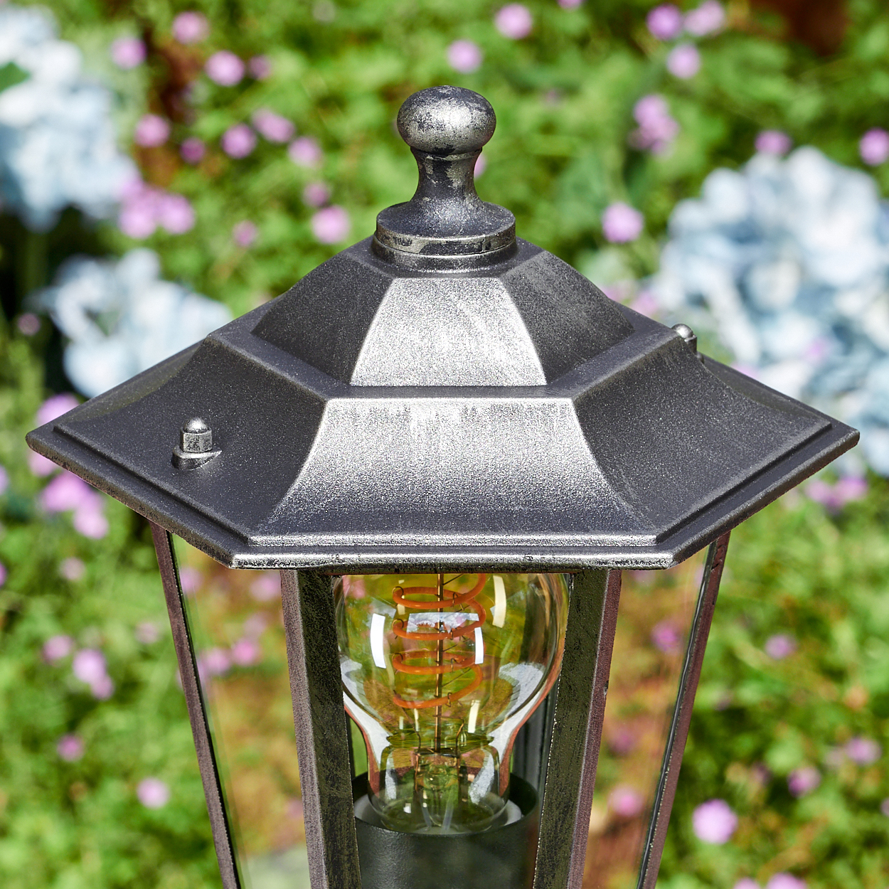 Nahaufnahme einer dekorativen Außenlampe mit Glühbirne. Die Lampe ist schwarz mit Metallic-Finish, vor verschwommenen Blumen.