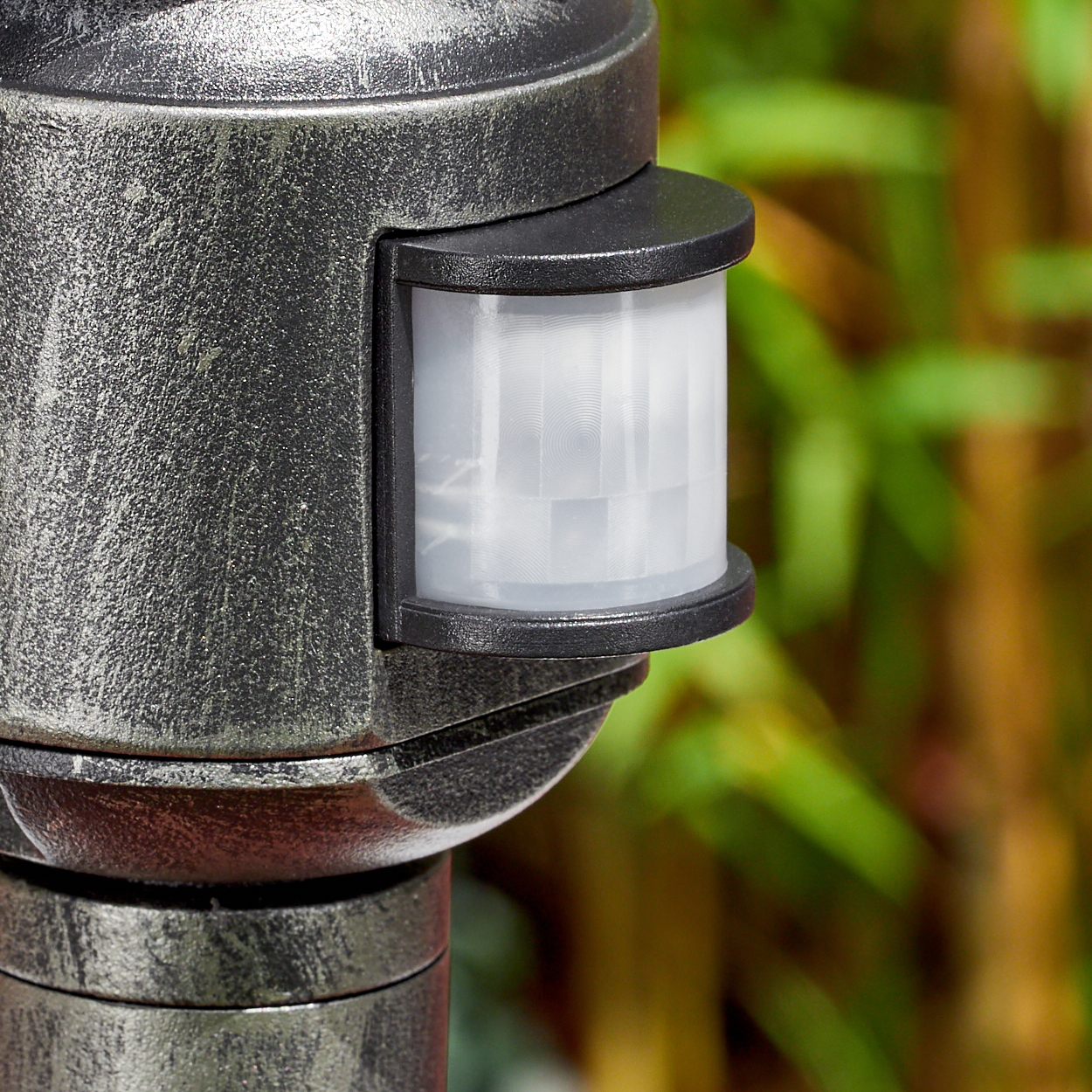 Nahaufnahme eines Bewegungssensors an einem Metallpfosten. Der Sensor ist weiß und schwarz, mit grünem Laub im Hintergrund.