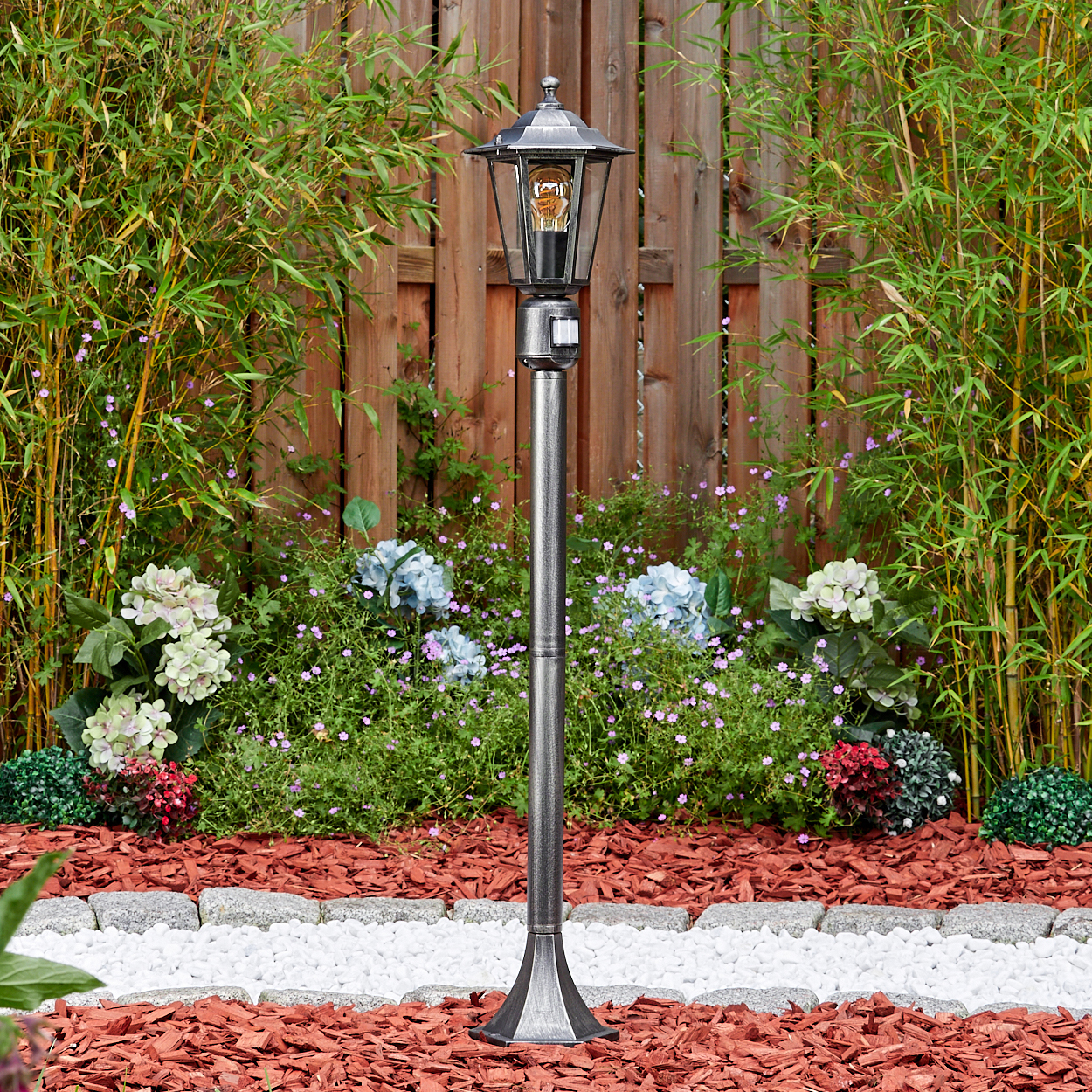 Eine graue Außenlampe steht in einem Garten. Im Hintergrund befinden sich Bambus, Blumen und ein Holzzaun.