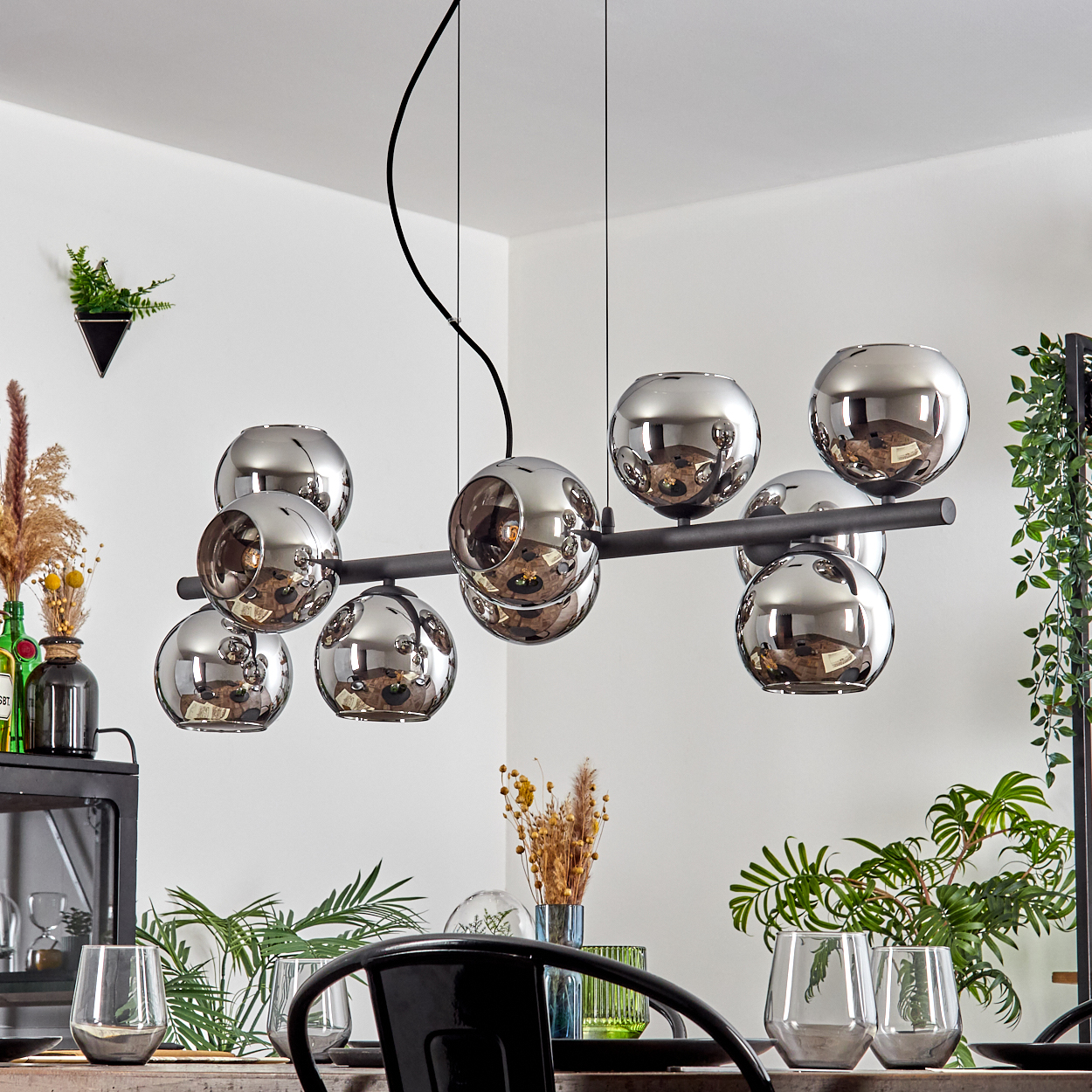 Ein modernes Esszimmer mit einem schwarzen Kronleuchter mit Glaskugeln. Pflanzen und Gläser stehen auf dem Tisch.