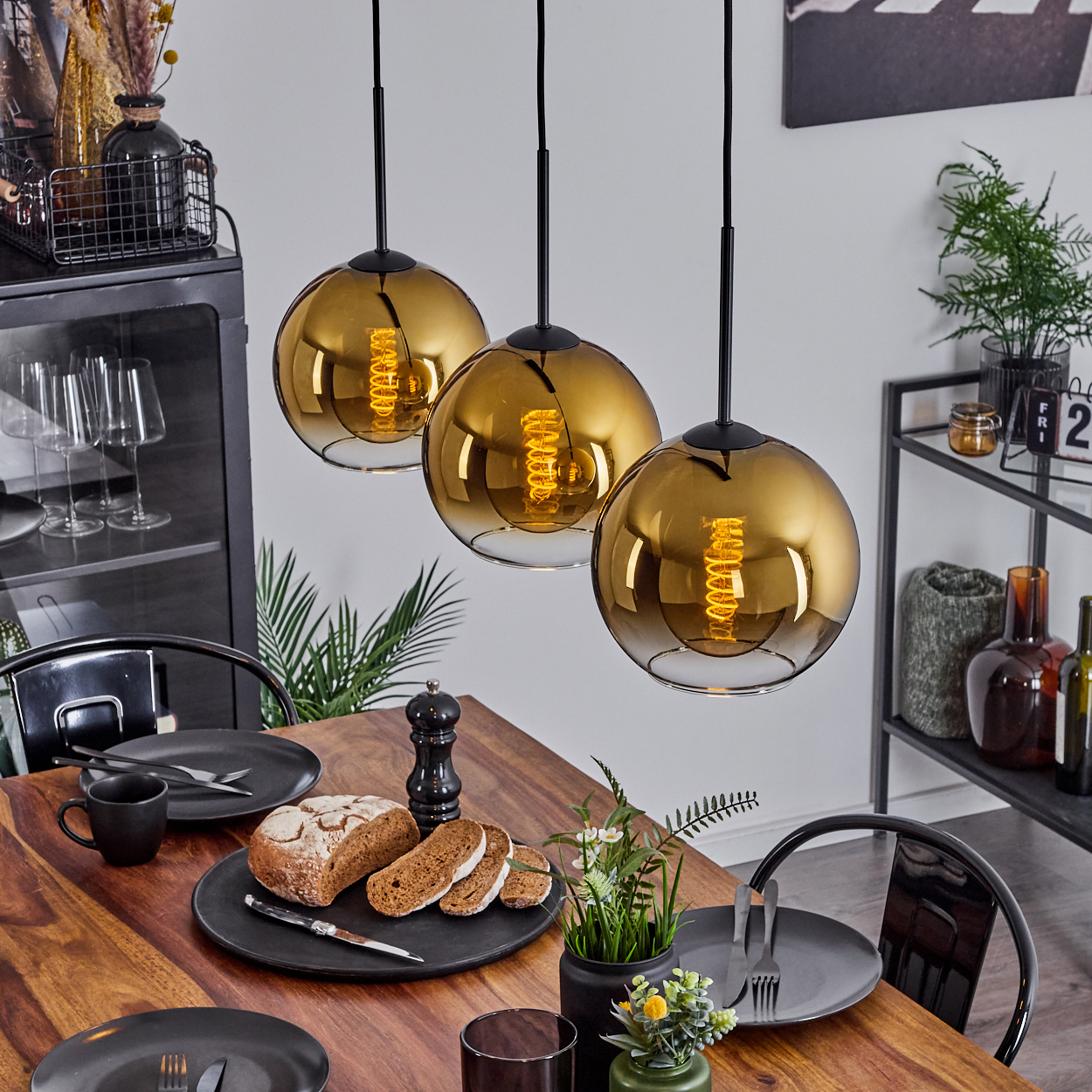 Esstisch mit Brot, Tellern und drei goldenen Pendelleuchten. Im Hintergrund ein schwarzer Schrank und Regal.