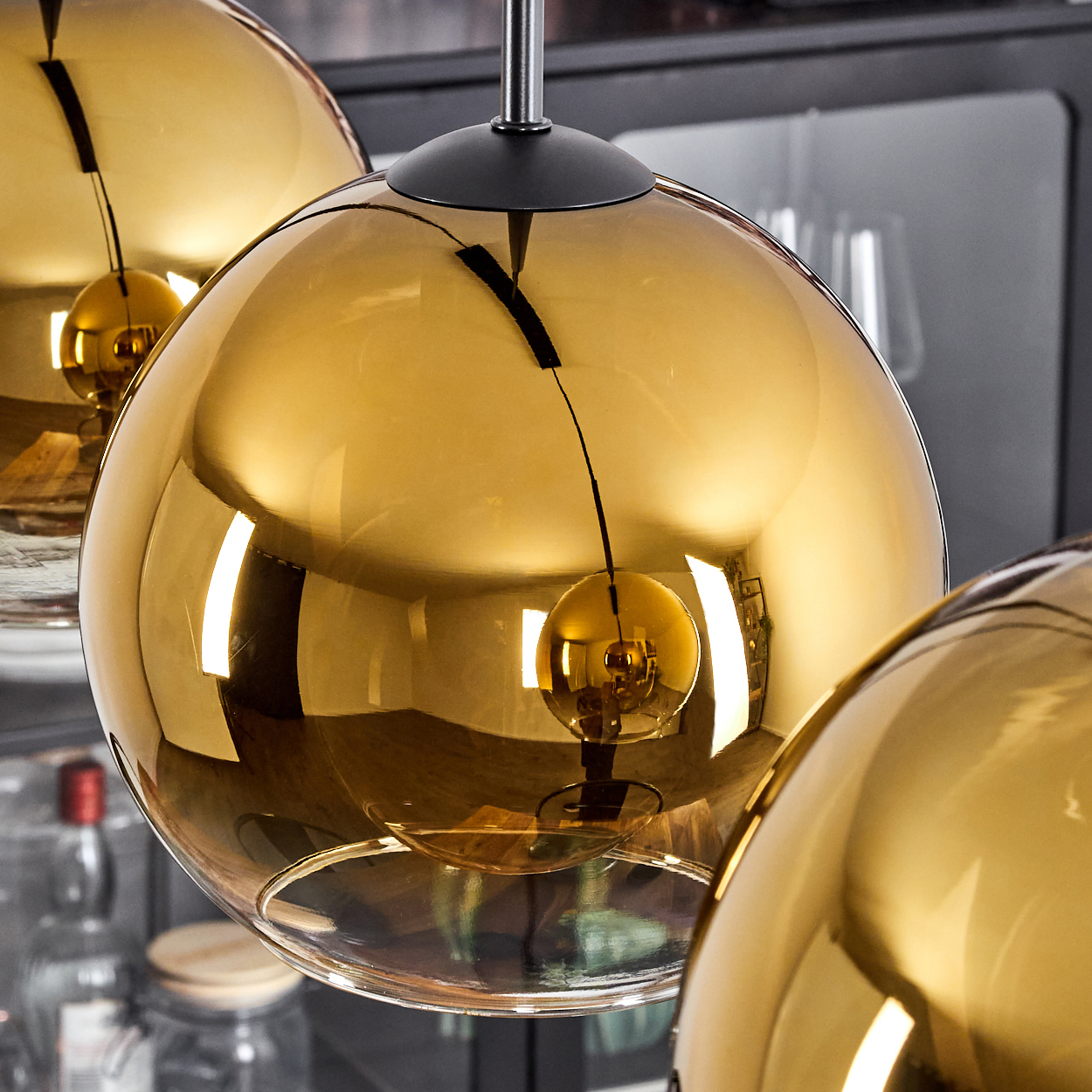 Nahaufnahme von drei goldenen, kugelförmigen Pendelleuchten. Jede reflektiert eine Glühbirne. Hintergrund verschwommene Regale.
