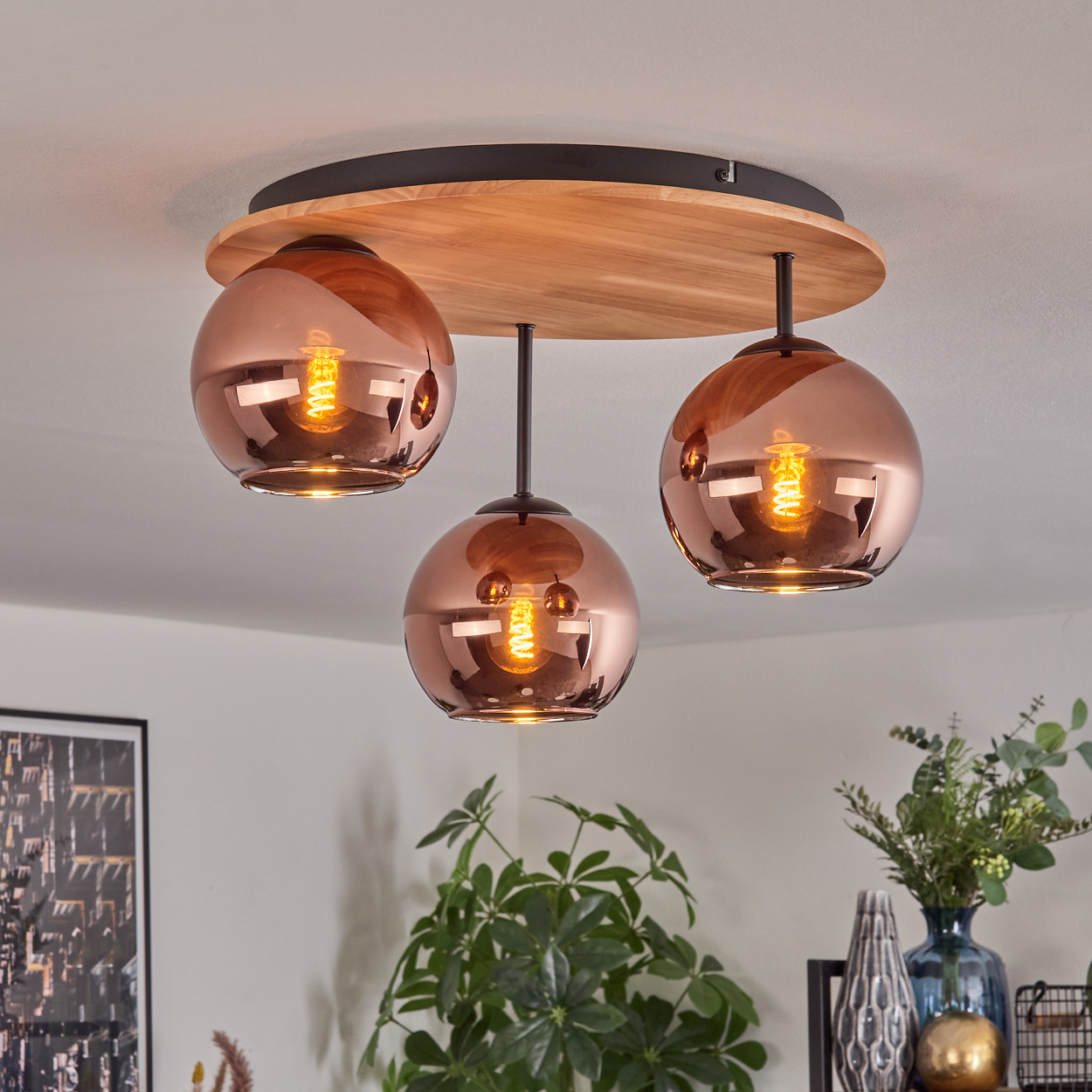 Eine moderne Deckenlampe mit drei kupferfarbenen Kugelleuchten, Holzsockel und einem schwarzen Ring. Pflanzen und Vasen im Hintergrund.
