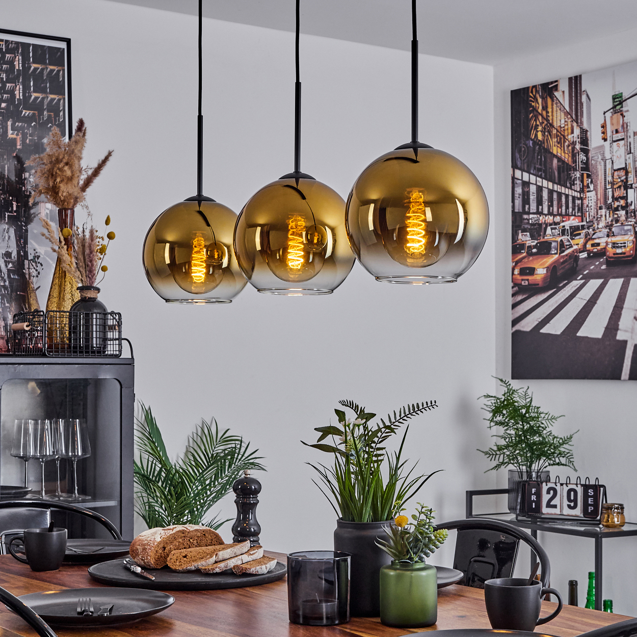 Drei goldene Pendelleuchten über einem Esstisch mit Tellern, Brot und Pflanzen. Eine New Yorker Stadtlandschaft dekoriert die Wand.
