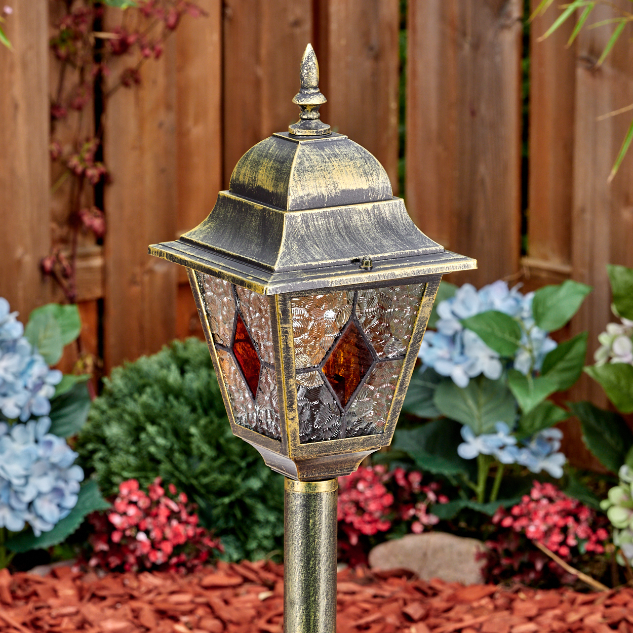 Eine verzierte Außenlampe mit Glasmalereien, in einem Garten mit Blumen und einem Holzzaun im Hintergrund.