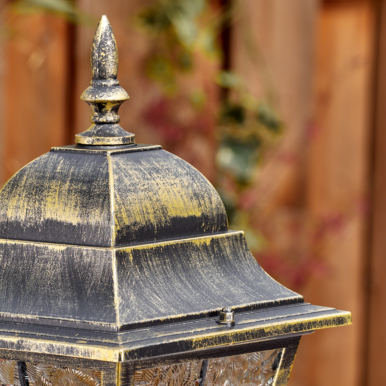 Nahaufnahme einer dekorativen Außenlampe. Sie hat eine schwarz-goldene Oberfläche. Der Hintergrund zeigt einen Holzzaun und etwas Laub.