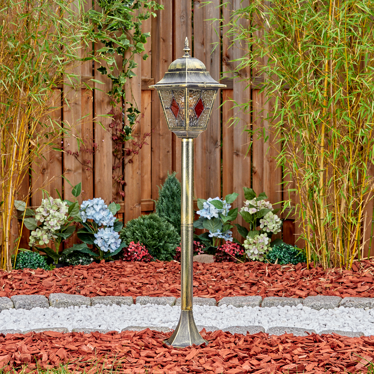 Eine dekorative Gartenlampe steht vor einem Holzzaun, umgeben von Pflanzen und roter Mulche.