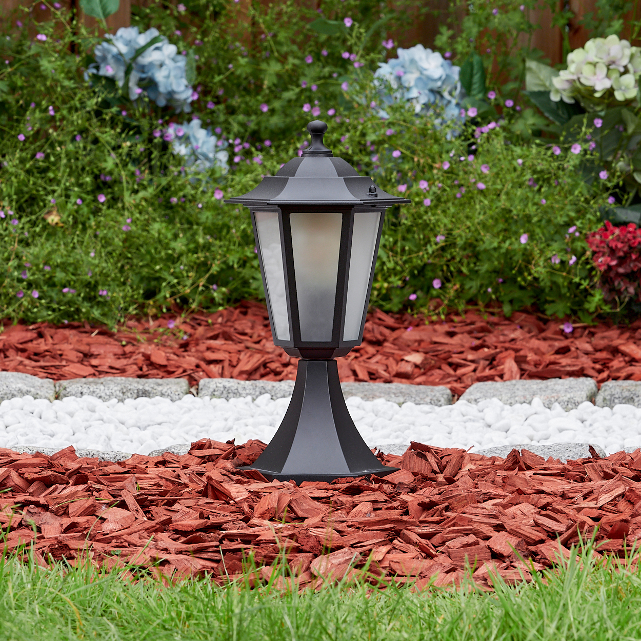 Eine schwarze Außenlampe steht in einem Beet mit roter Mulche und weißen Steinen, mit Blumen und grünem Gras im Hintergrund.