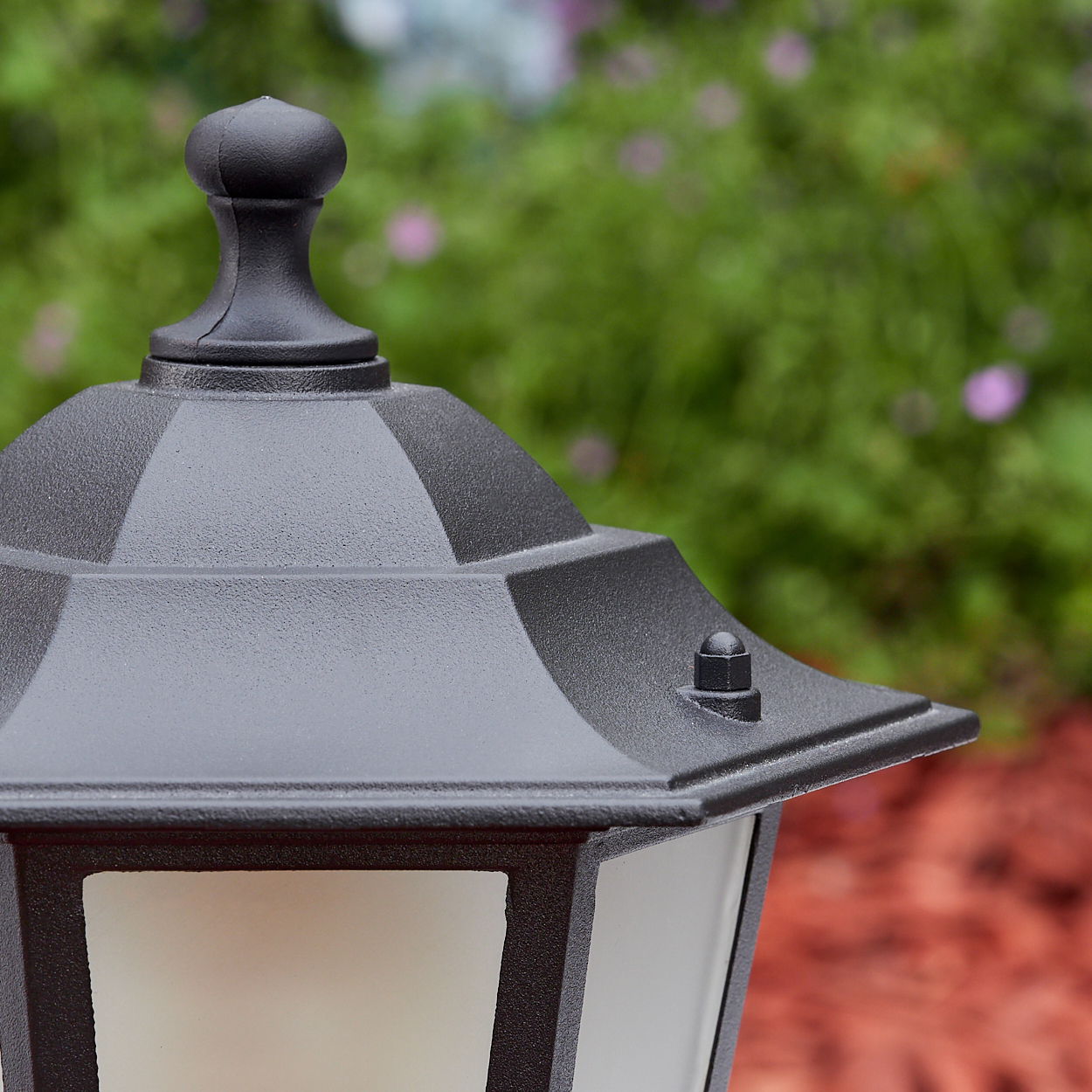 Nahaufnahme einer schwarzen Außenlampe. Die Lampe hat eine dekorative Oberseite und ein weißes Milchglas. Grünes Laub im Hintergrund.
