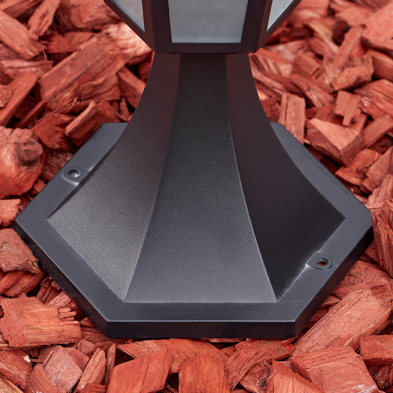 Ein schwarzer Lampensockel steht auf rotem Mulch. Die Lampe hat ein geometrisches Design und besteht aus Metall.