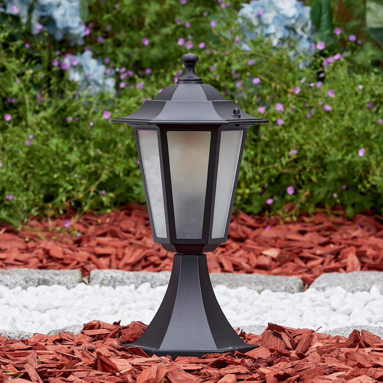 Eine schwarze Außenlampe steht auf roter Mulch und weißen Steinen, mit einem grünen Busch und Blumen im Hintergrund.