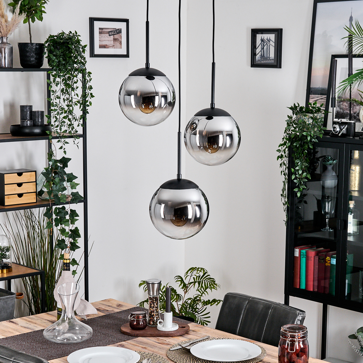 Esszimmer mit Holztisch, drei hängenden Kugelleuchten, Pflanzen und gerahmten Bildern. Teller und ein Glas stehen auf dem Tisch.