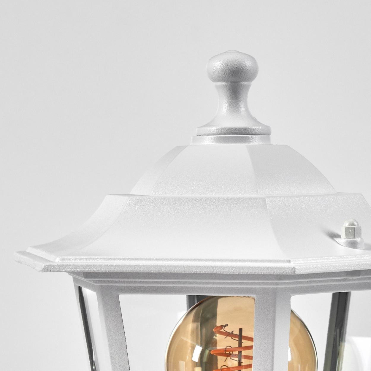 Nahaufnahme einer weißen Außenlampe. Die Lampe hat eine dekorative Oberseite, Glaspaneele und eine sichtbare Glühbirne.