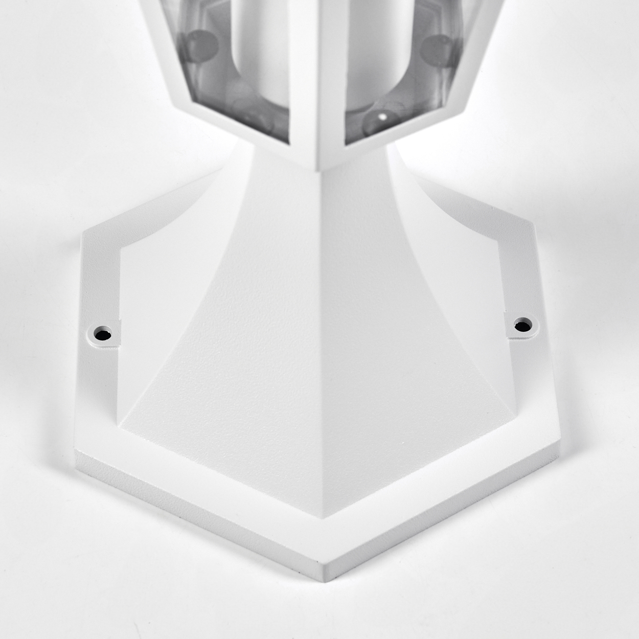Eine weiße, geometrische Außenlampe. Sie hat einen sechseckigen Sockel mit Befestigungslöchern und einen transparenten oberen Bereich.