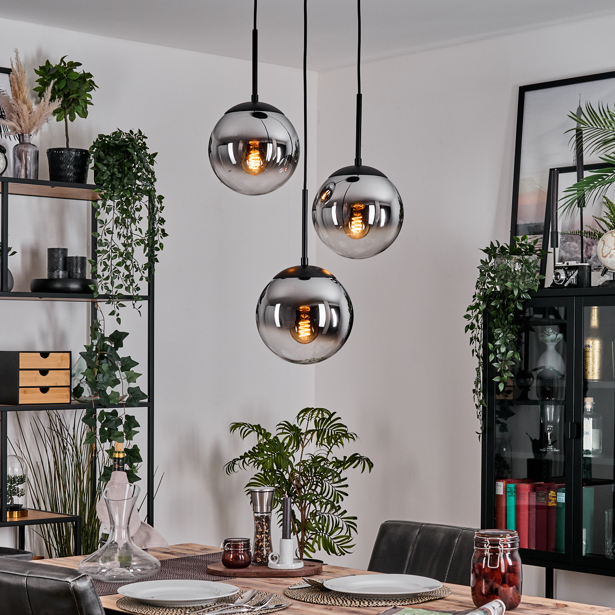 Esszimmer mit drei kugelförmigen Pendelleuchten. Tisch mit Tellern gedeckt. Regale mit Pflanzen und Dekor. Schwarzer Schrank.