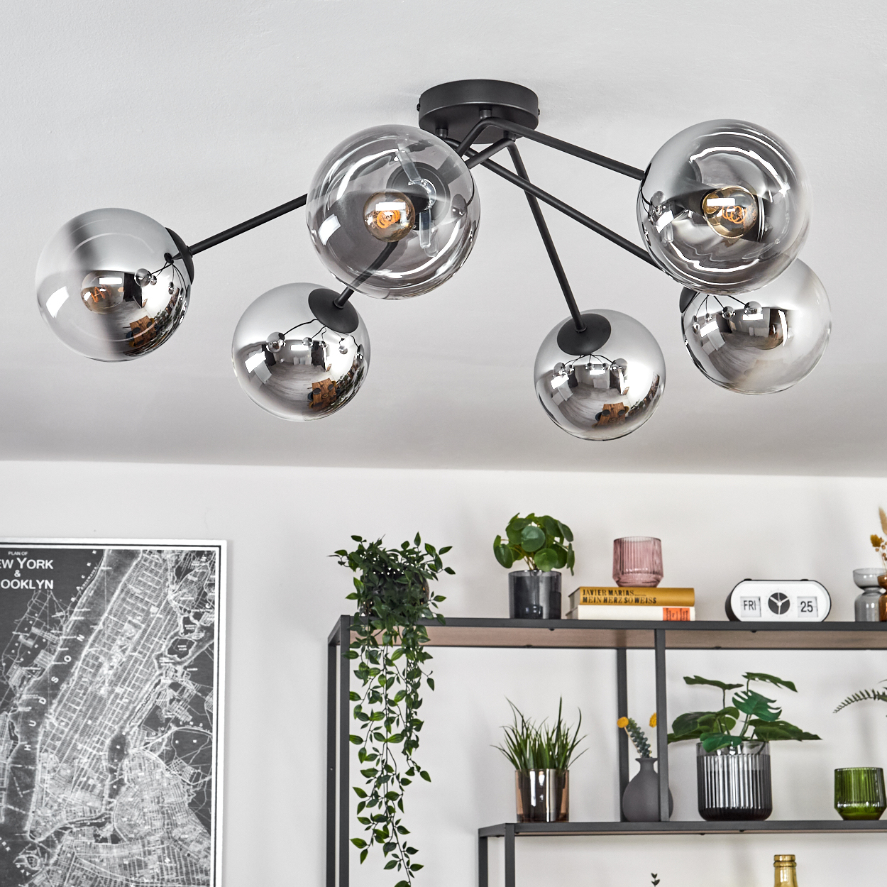 Moderne Deckenleuchte mit sechs kugelförmigen Glasschirmen. Darunter ein Regal mit Pflanzen, Büchern und Dekoration. An der Wand hängt eine Karte.