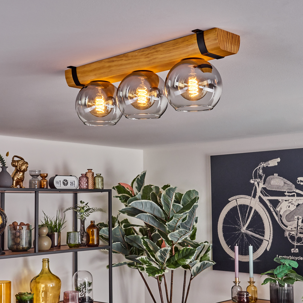 Deckenlampe mit drei Glühbirnen, Holzbalken, Glaskugeln. Regale mit Dekoration und eine große Pflanze. Ein Bild eines Motorrads an der Wand.