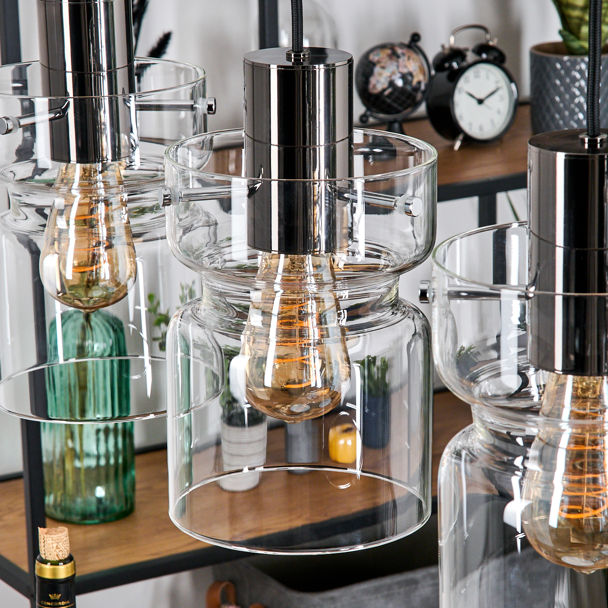 Nahaufnahme von Glas-Pendelleuchten mit Edison-Glühbirnen. Im Hintergrund ein Regal mit Uhr, Globus und Pflanzen.