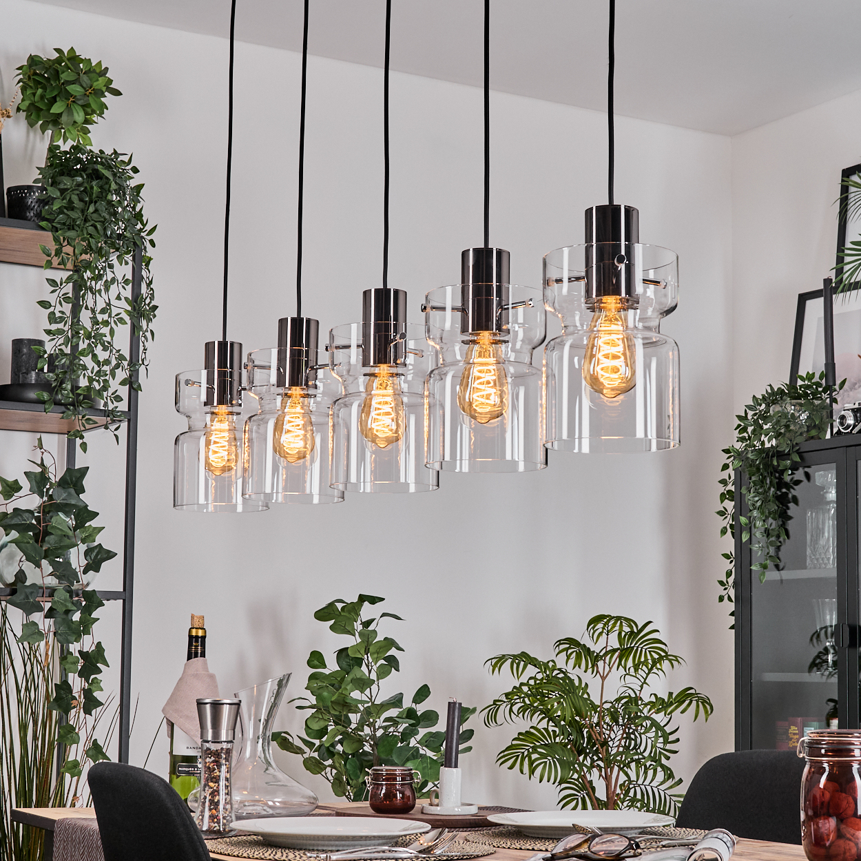 Esszimmer mit Hängelampen. Die Lampen haben Glasschirme. Im Hintergrund sind Pflanzen, ein Tisch, eine Weinflasche und ein Schrank.