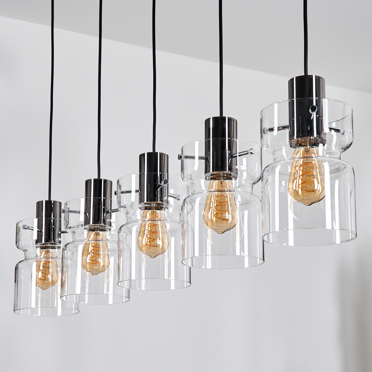 Sechs Pendelleuchten mit klaren Glasschirmen und Edison-Glühbirnen hängen an schwarzen Kabeln vor einer weißen Wand.