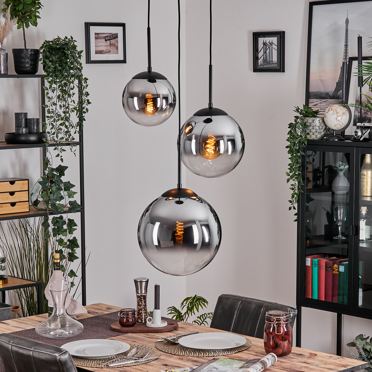 Drei kugelförmige Pendelleuchten hängen über einem Holzesstisch, der für eine Mahlzeit gedeckt ist. Der Hintergrund umfasst Regale, einen Schrank und gerahmte Kunst.