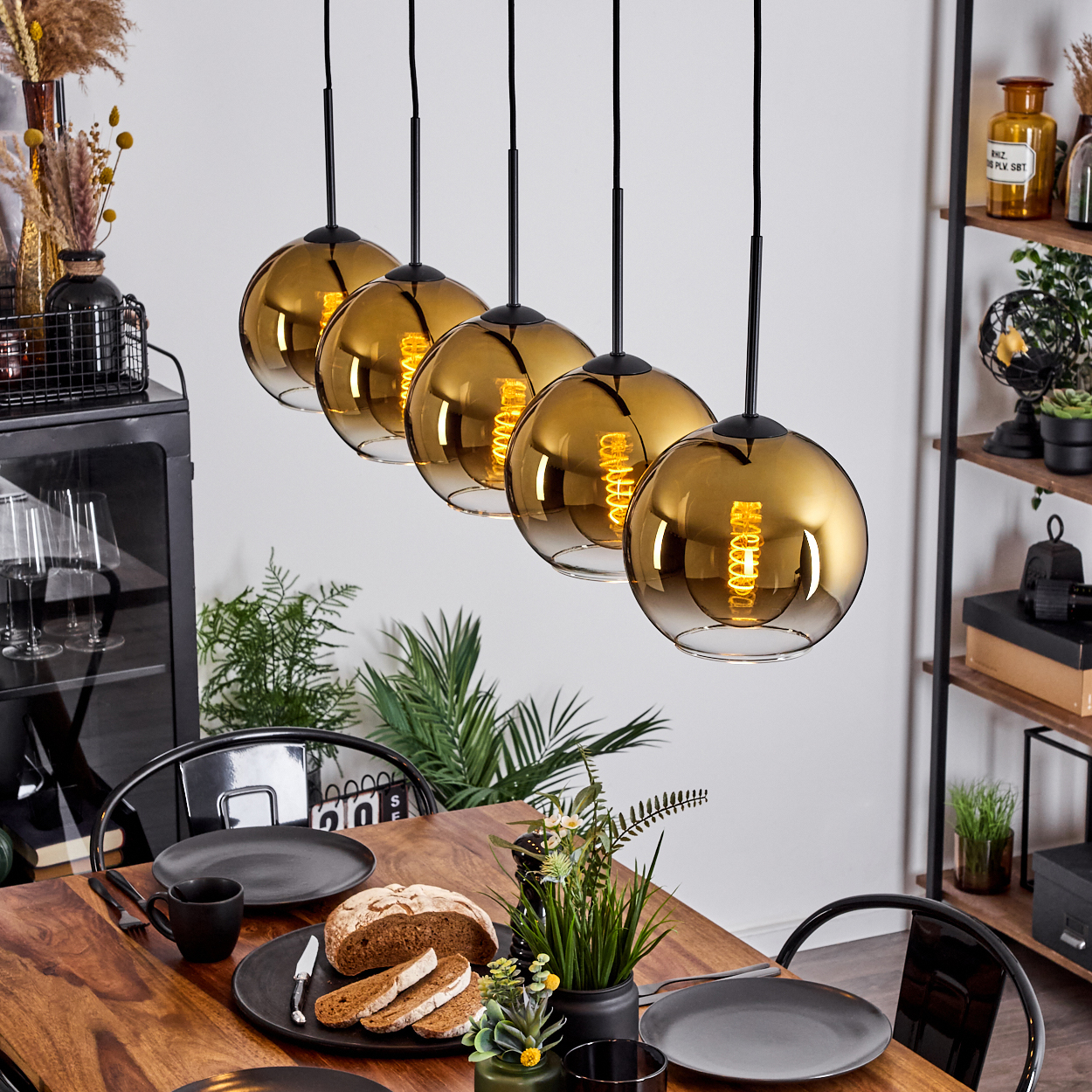 Esszimmer mit Holztisch, schwarzen Stühlen und fünf hängenden Kugelleuchten. Tisch gedeckt mit Tellern, Brot und Pflanzen.