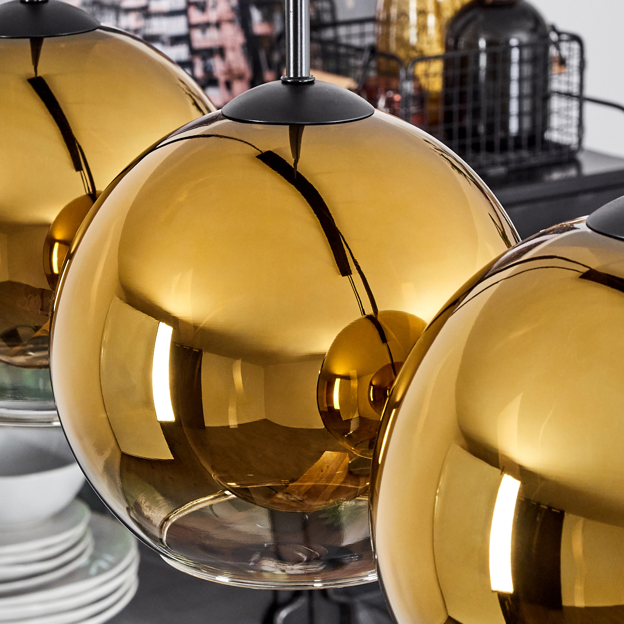Drei goldene, kugelförmige Pendelleuchten. Jede hat eine schwarze Kappe. Der Hintergrund umfasst einen Metallkorb und Geschirr.
