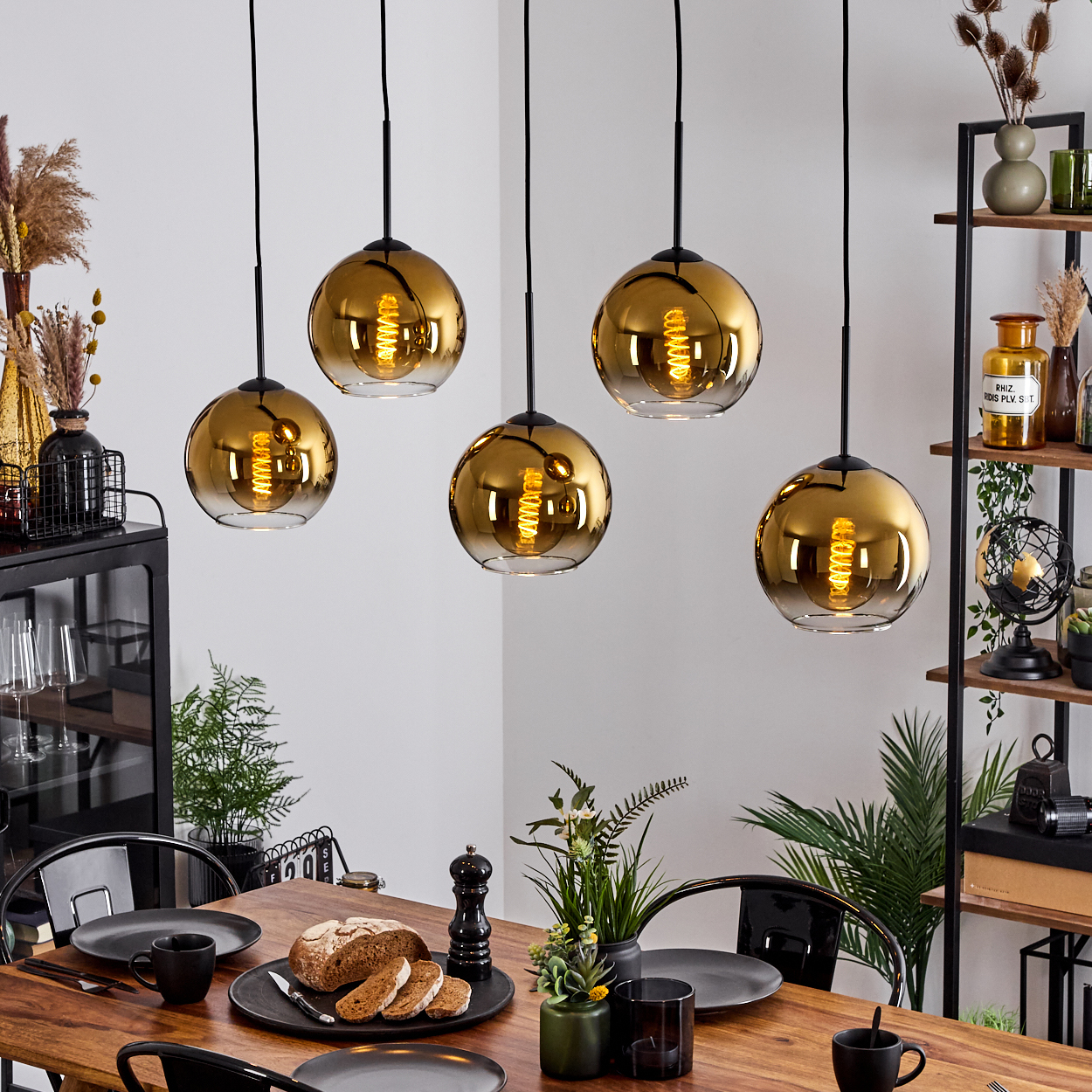 Esszimmer mit Holztisch und gedecktem Tisch. Fünf goldene Pendelleuchten hängen darüber. Brot und Teller sind sichtbar.