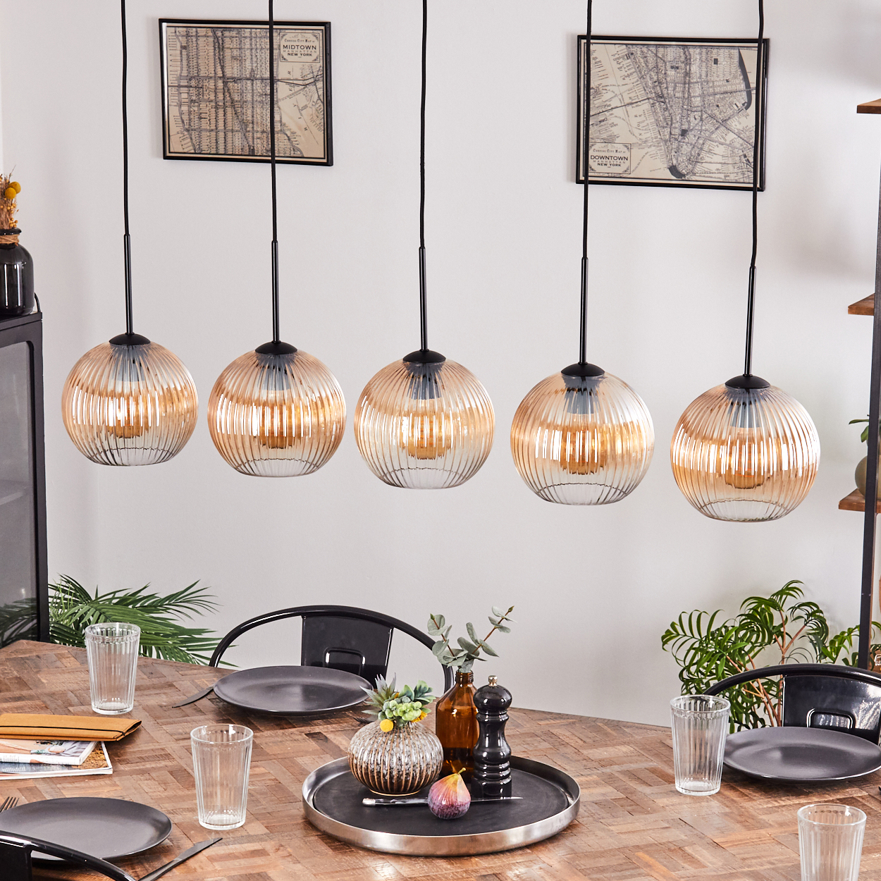 Esszimmer mit Holztisch, fünf Hängelampen, Tellern, Gläsern und einem dekorativen Tablett mit einer Feige.