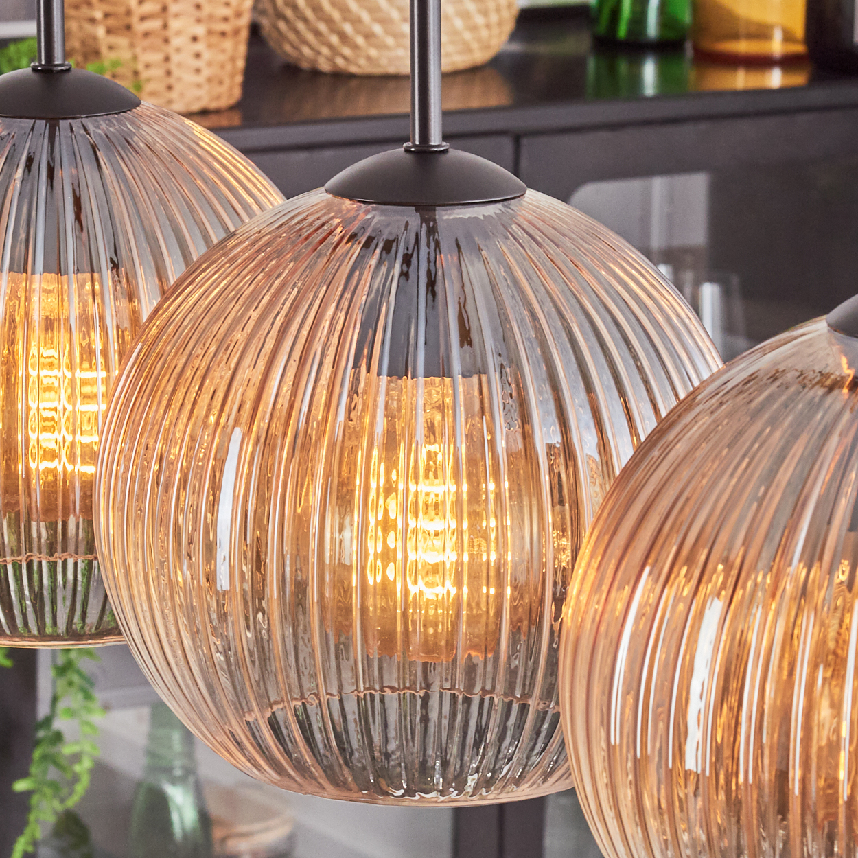 Drei beleuchtete Pendelleuchten aus Glas mit geripptem Design. Warmes Licht scheint im Inneren. Im Hintergrund sind Weidenkörbe und dunkle Möbel zu sehen.
