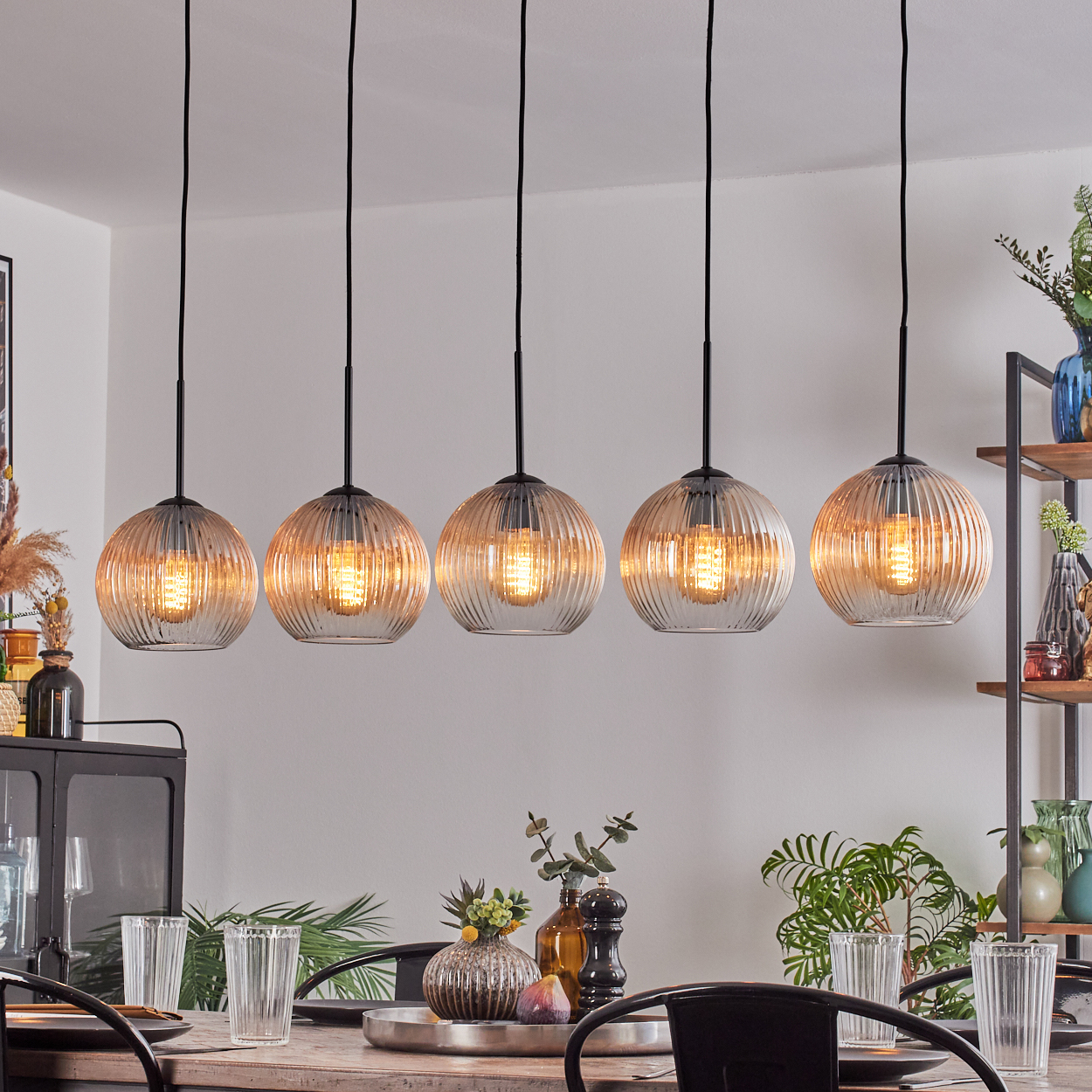Fünf Pendelleuchten über einem Esstisch. Die Lampen sind aus Glas mit Glühbirnen. Tisch mit Gläsern und Tellern.