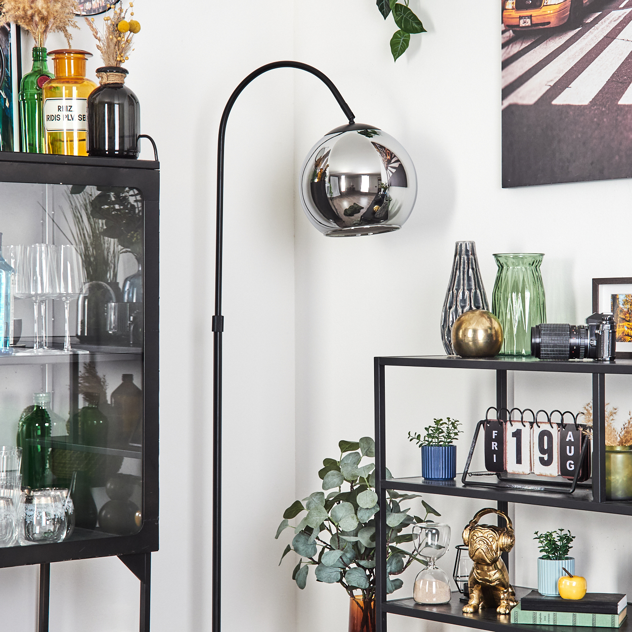 Ein Raum mit Stehlampe, Regalen mit Vasen, einer Kamera und einem Kalender. Ein schwarzer Schrank zeigt Glaswaren und Flaschen.