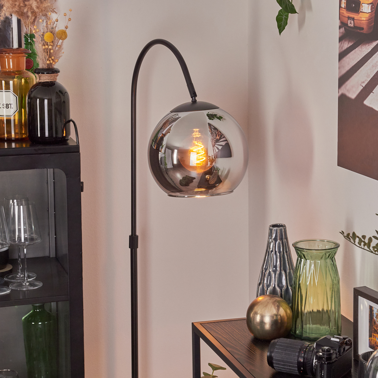 Eine moderne Stehlampe mit kugelförmigem Glasschirm, die ein warmes Licht ausstrahlt. Verschiedene Dekorationsartikel sind darum platziert.