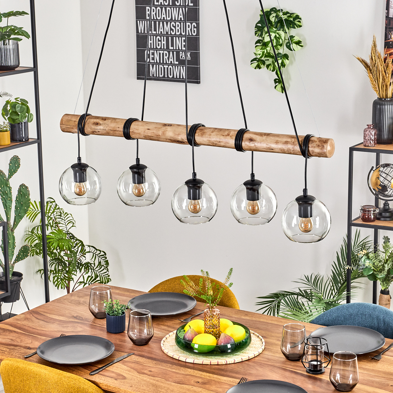 Esstisch mit Tellern, Gläsern und einer Obstschale. Über dem Tisch hängt ein Holzbalken mit Kugellampen. Pflanzen und Dekor.