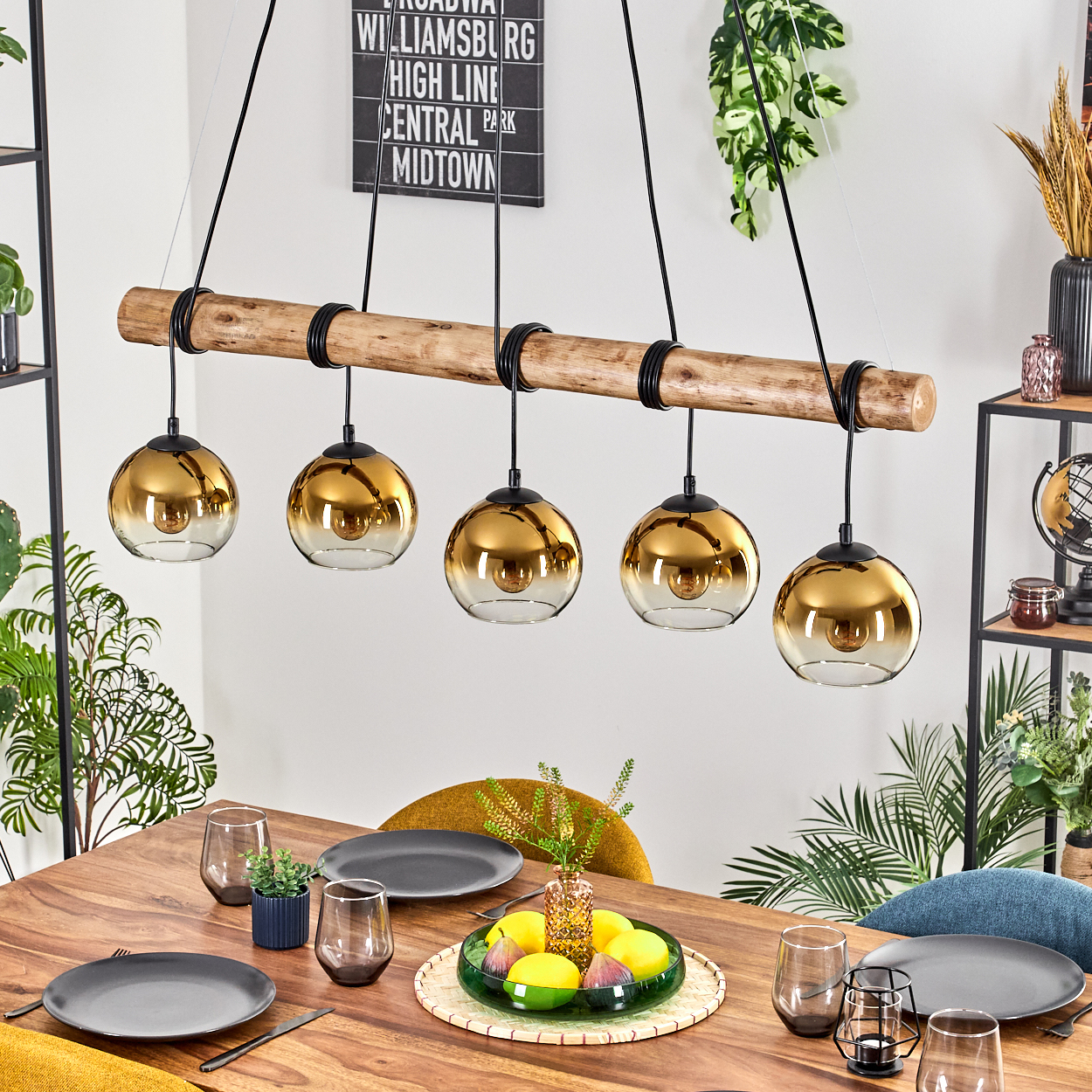 Esstisch mit Holzplatte, verziert mit Tellern, Gläsern und einer Obstschale. Darüber hängen fünf goldene Pendelleuchten.