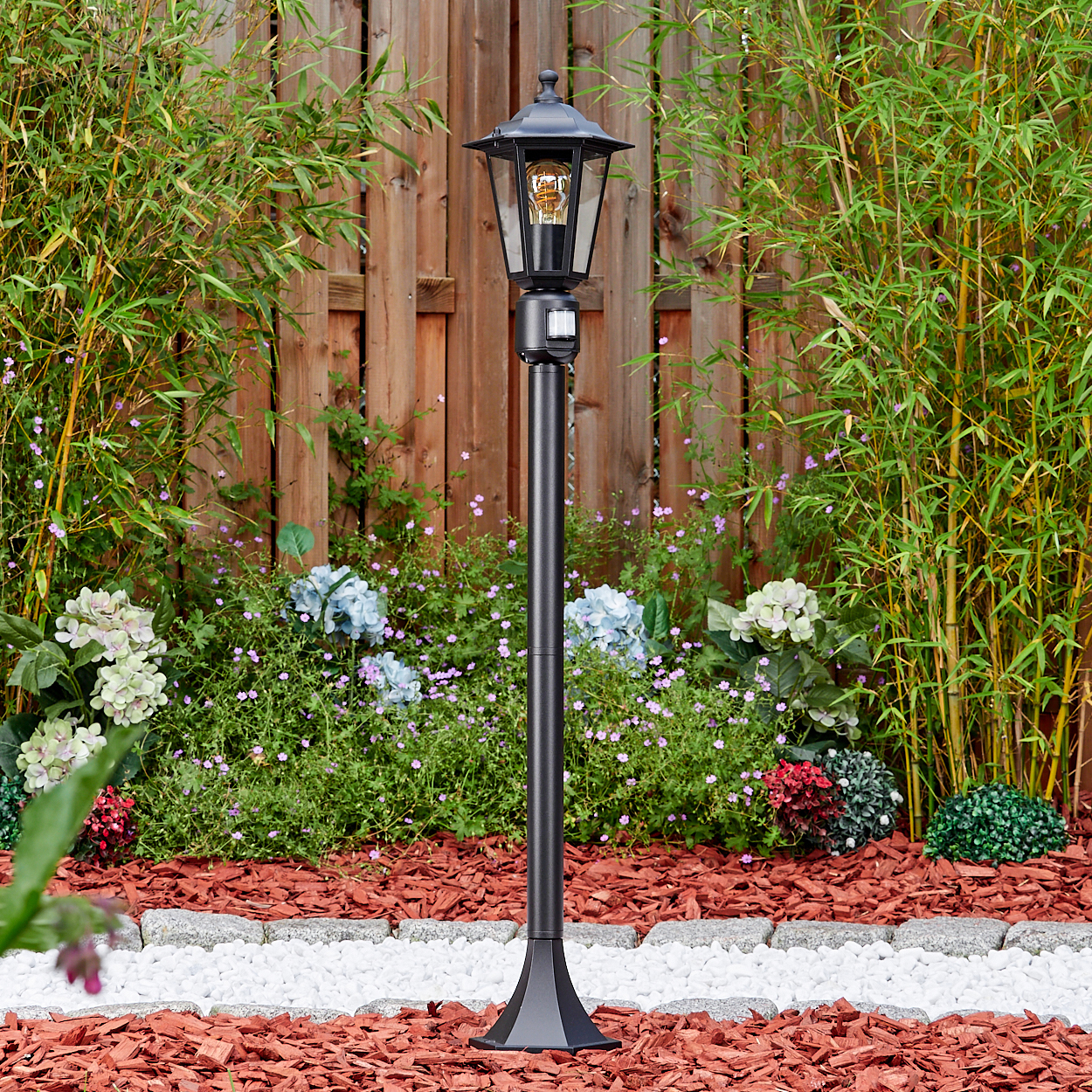 Eine schwarze Außenlampe steht in einem Garten. Bambus, Blumen und ein Holzzaun sind im Hintergrund. Rindenmulch und weiße Steine sind im Vordergrund.