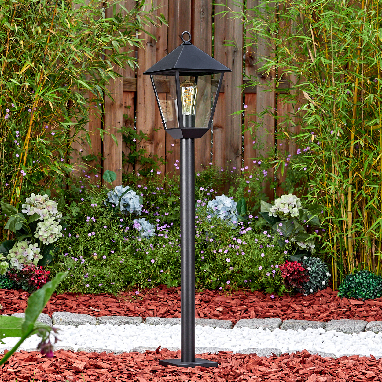 Eine schwarze Außenlampe steht in einem Garten mit Blumen und Bambus. Rote Mulch und weiße Steine säumen die Blumenbeete. Holzzaun im Hintergrund.